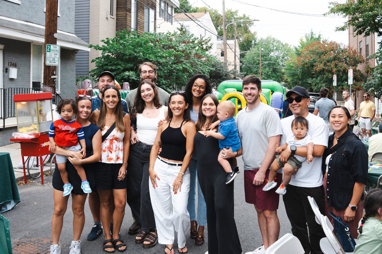 Neighbors and children pose together on a residential street beside a popcorn cart and inflatable play structure.