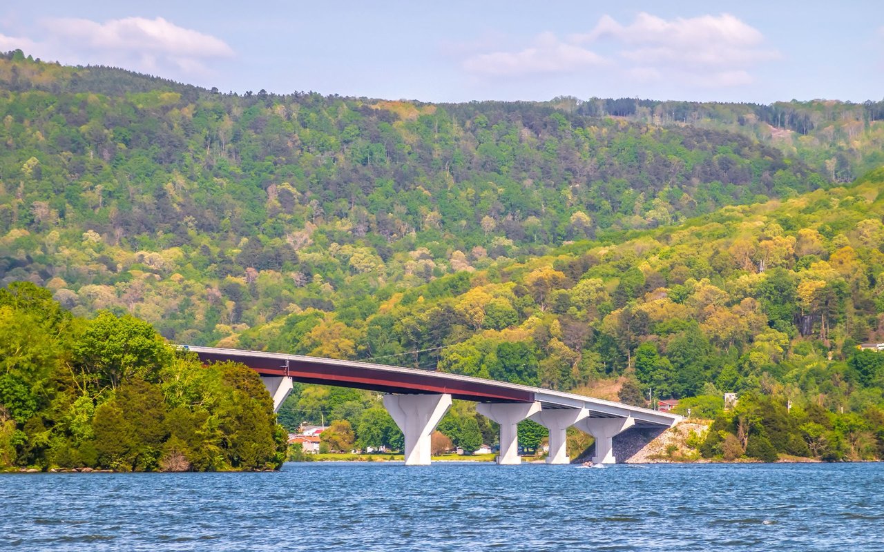 Bridge crossing a lake below wooded hills, with scattered shoreline buildings and partly cloudy skies.