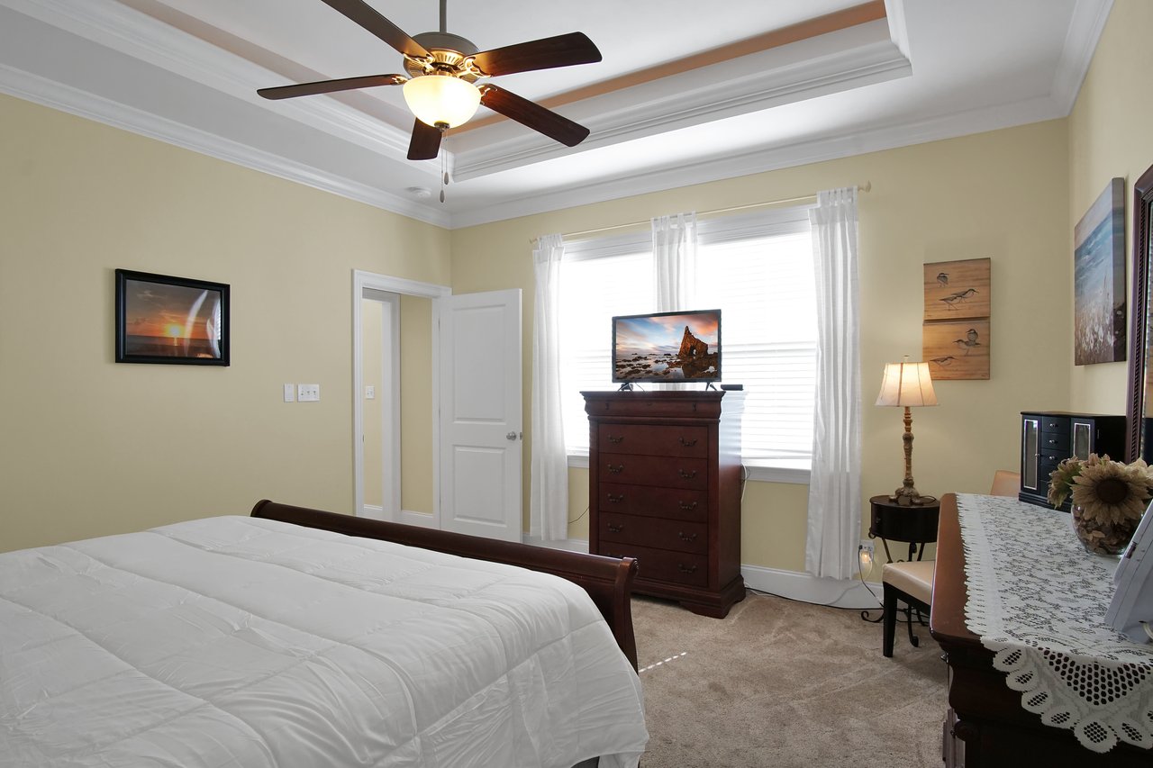 Cozy bedroom with a white bedspread, ceiling fan, and dark wood dresser. Sunlight filters through curtains, creating a warm and inviting atmosphere.