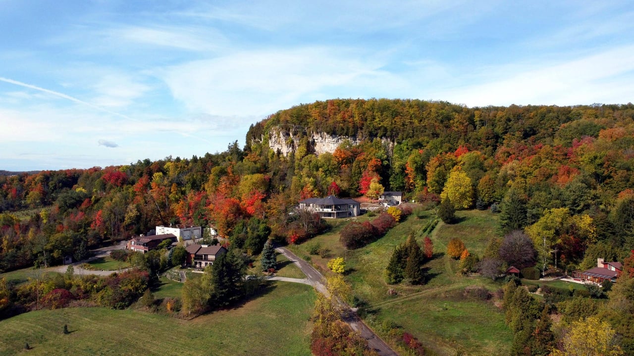An aerial drone photograph of custom luxury homes nestled at the base of a rocky cliff along the Niagara Escarpment during autumn, surrounded by vibrant red, orange, and yellow fall foliage, showcasing the natural landscape and real estate appeal of living in Milton Ontario.