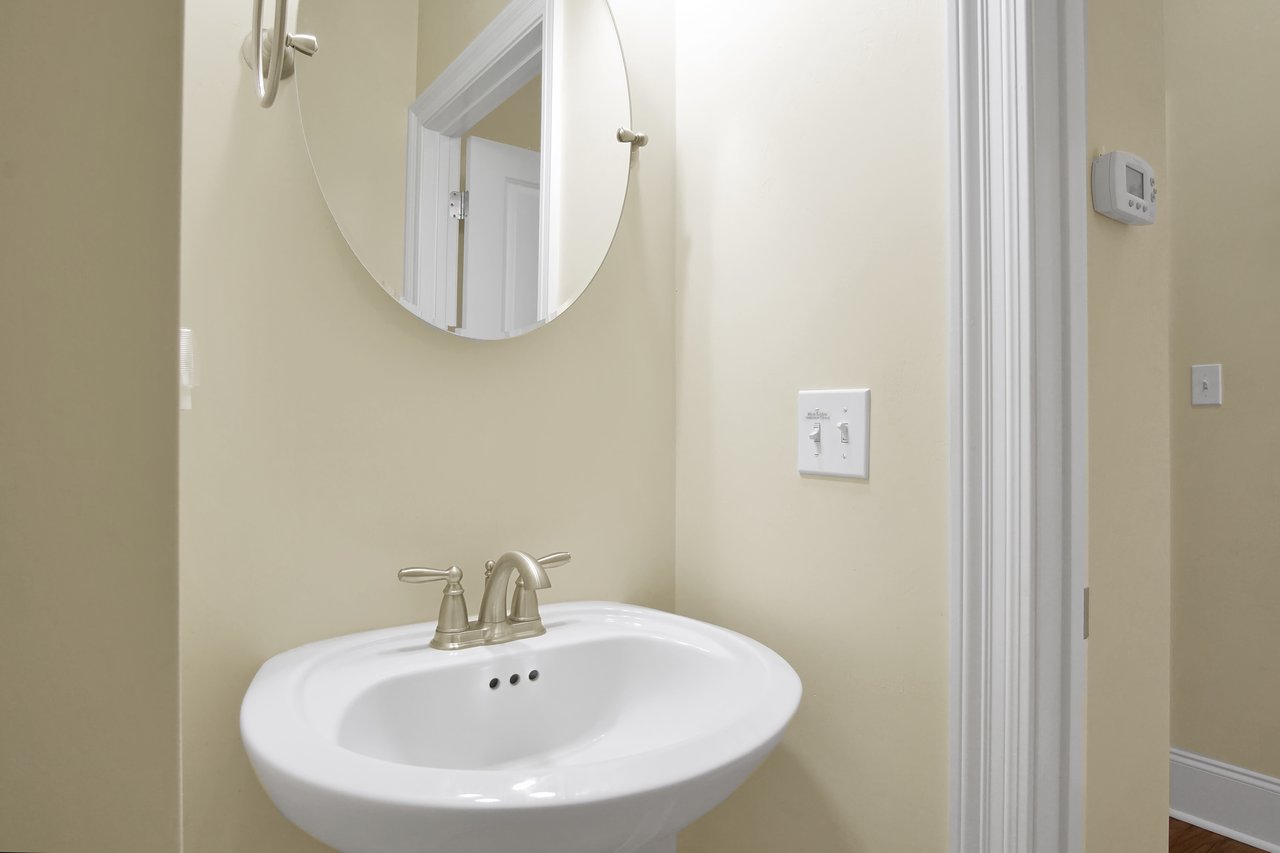 Small bathroom with a round mirror above a white pedestal sink, featuring sleek silver faucets. Light switch and thermostat on a cream-colored wall. Elegant and clean atmosphere.