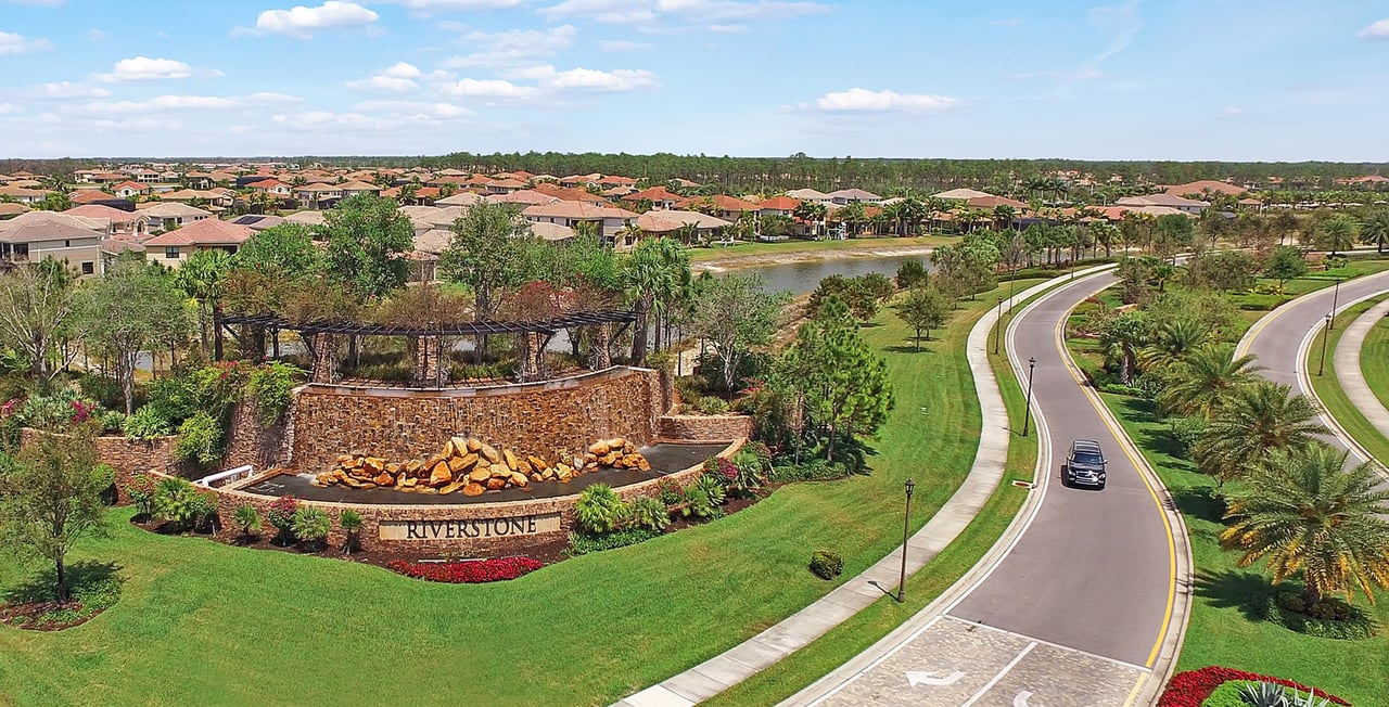 Riverstone neighborhood entrance with a stone monument sign, curving road, lake, and homes.