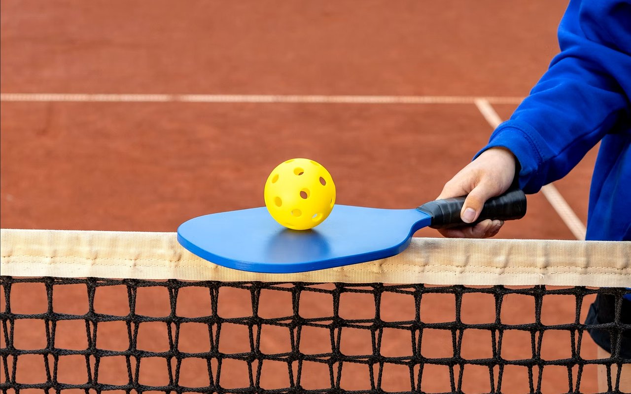 Blue pickleball paddle and yellow ball resting above a net on a reddish court.