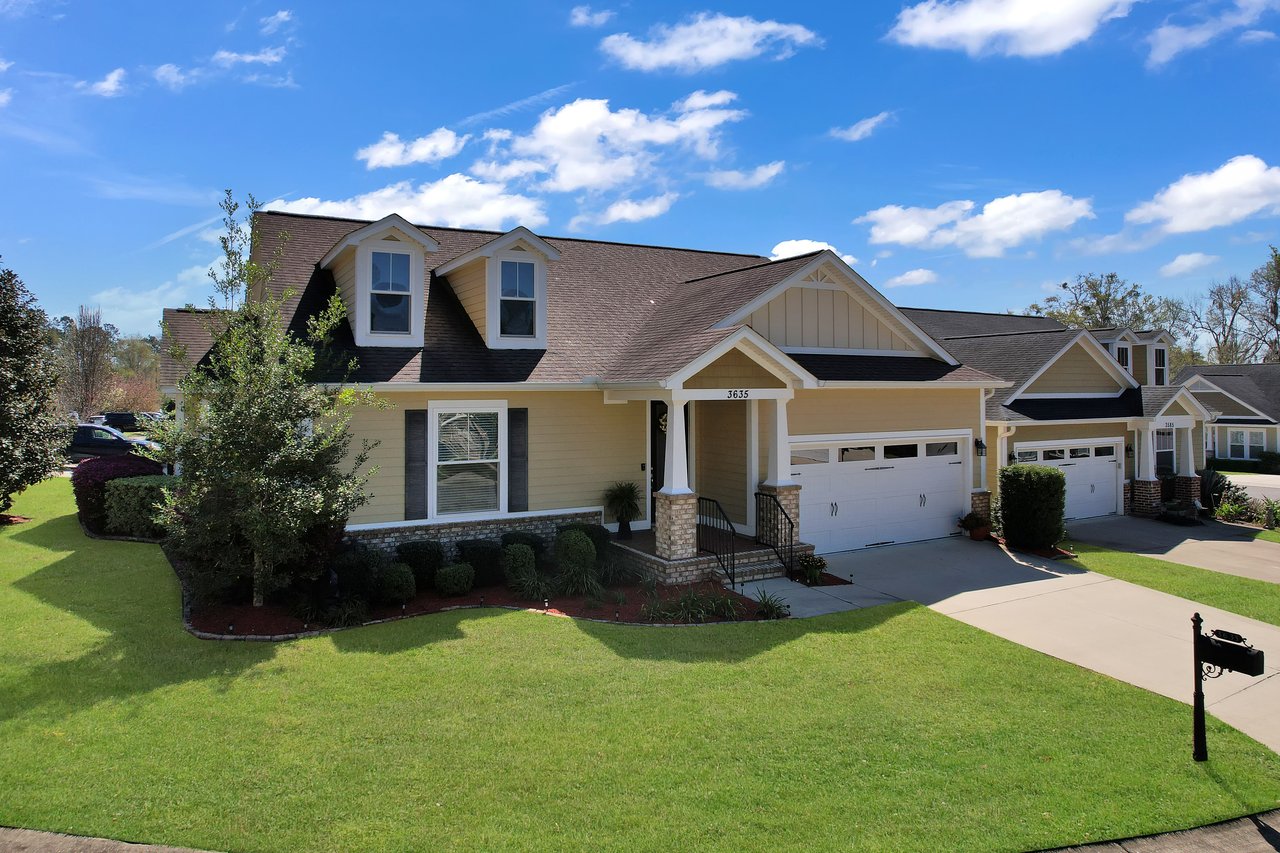 Charming suburban home with a manicured lawn under a blue sky. Features include a double garage, brick porch, and trees, evoking a peaceful neighborhood feel.