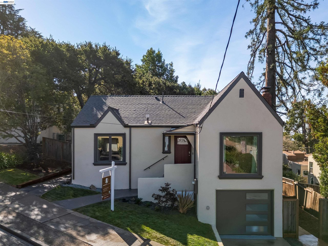 Gray stucco house with steep gables, dark trim, an attached garage, and a real estate sign.