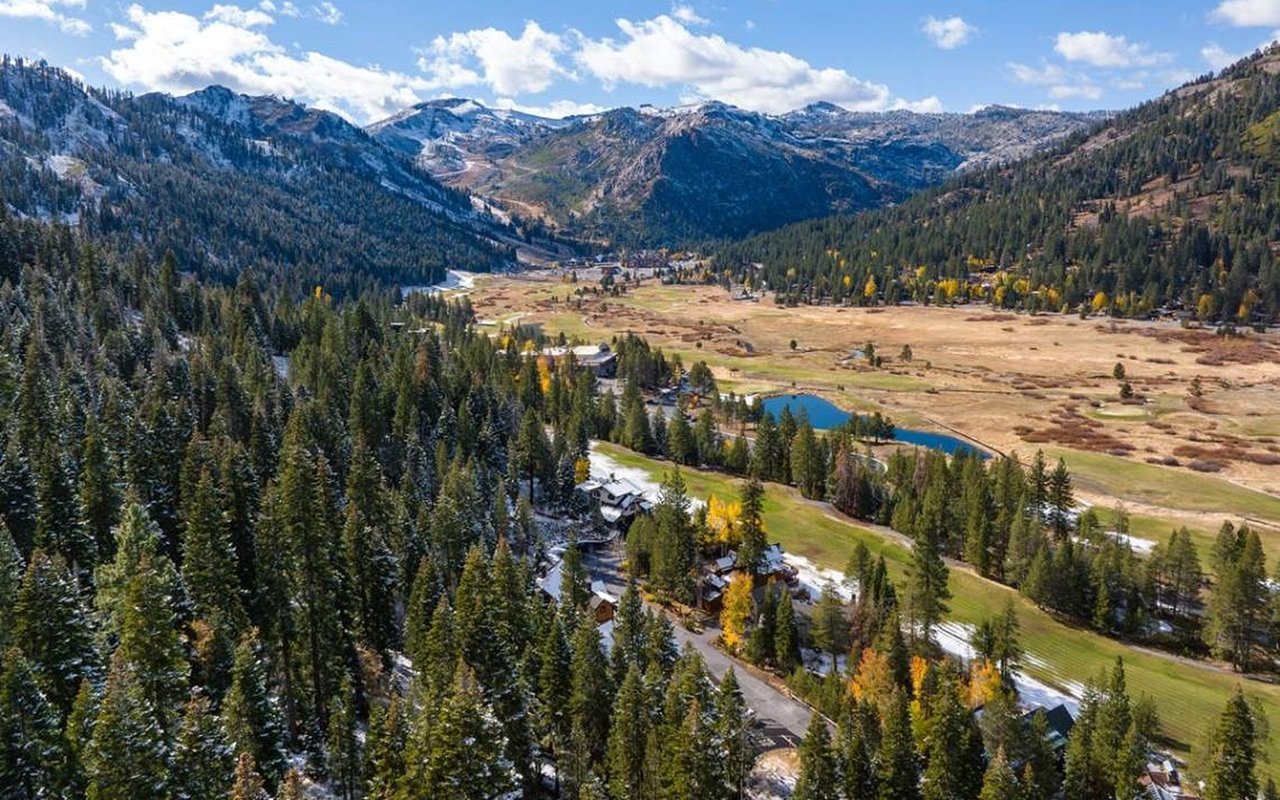 Aerial view of homes in a mountain valley, with a pond, autumn trees, and snow-dusted peaks.