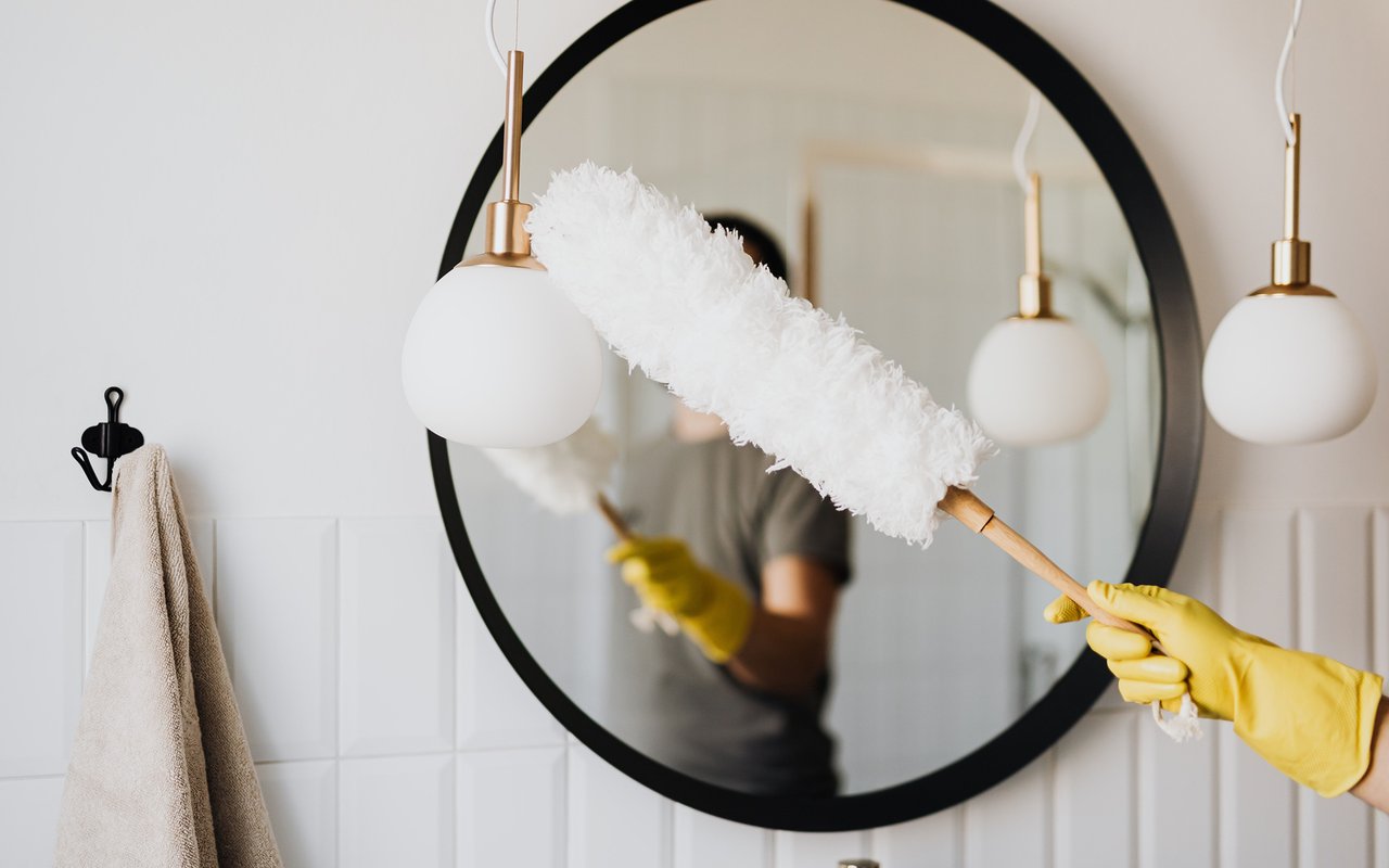 A yellow-gloved hand dusts a large round bathroom mirror with a white duster.
