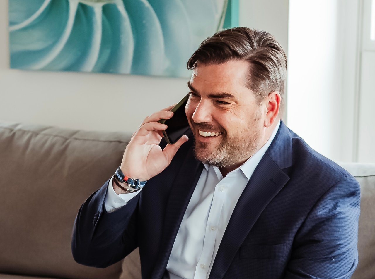 Smiling man in a navy suit talking on a smartphone from a sofa.