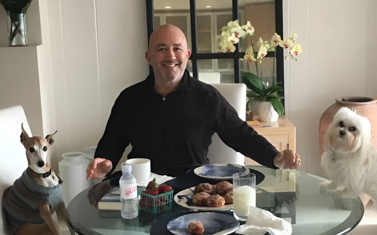A smiling man sits at breakfast with a sweater-wearing dog and a fluffy white dog.