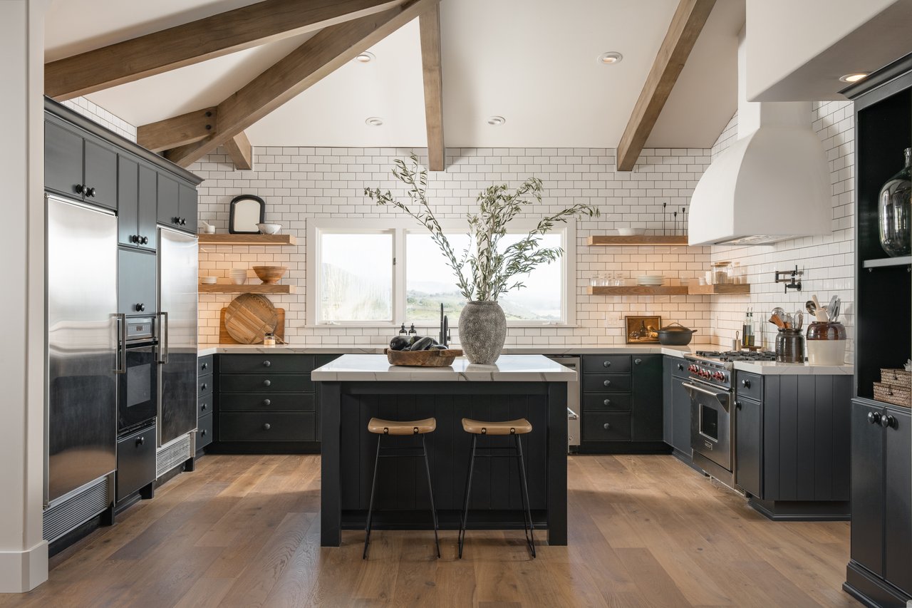 Kitchen with exposed wood beams, dark cabinets, and a central island beneath large windows.