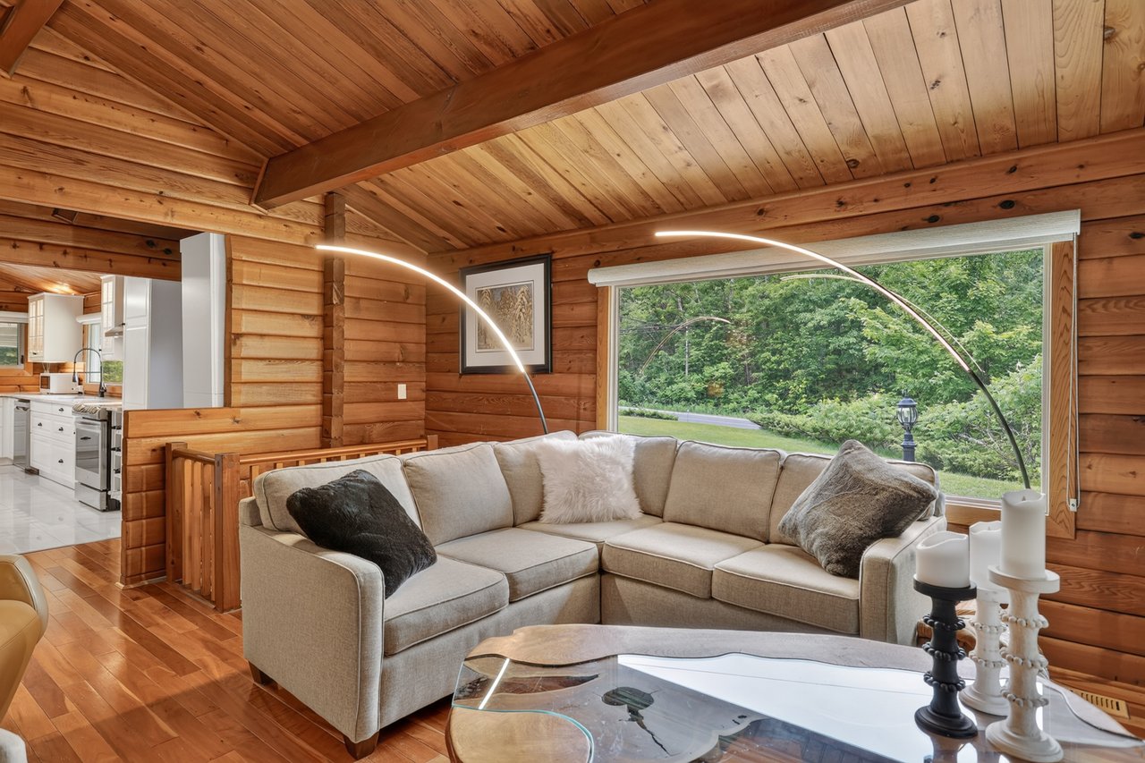 Wood-paneled living room with a beige sectional, vaulted timber ceiling, and large window overlooking greenery.