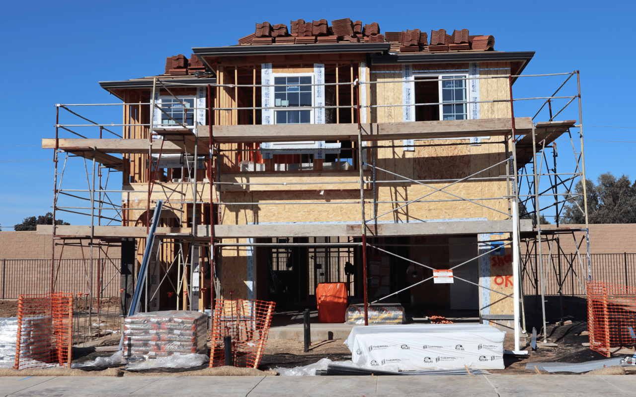 Two-story home under construction with scaffolding, exposed sheathing, and a partially tiled roof.