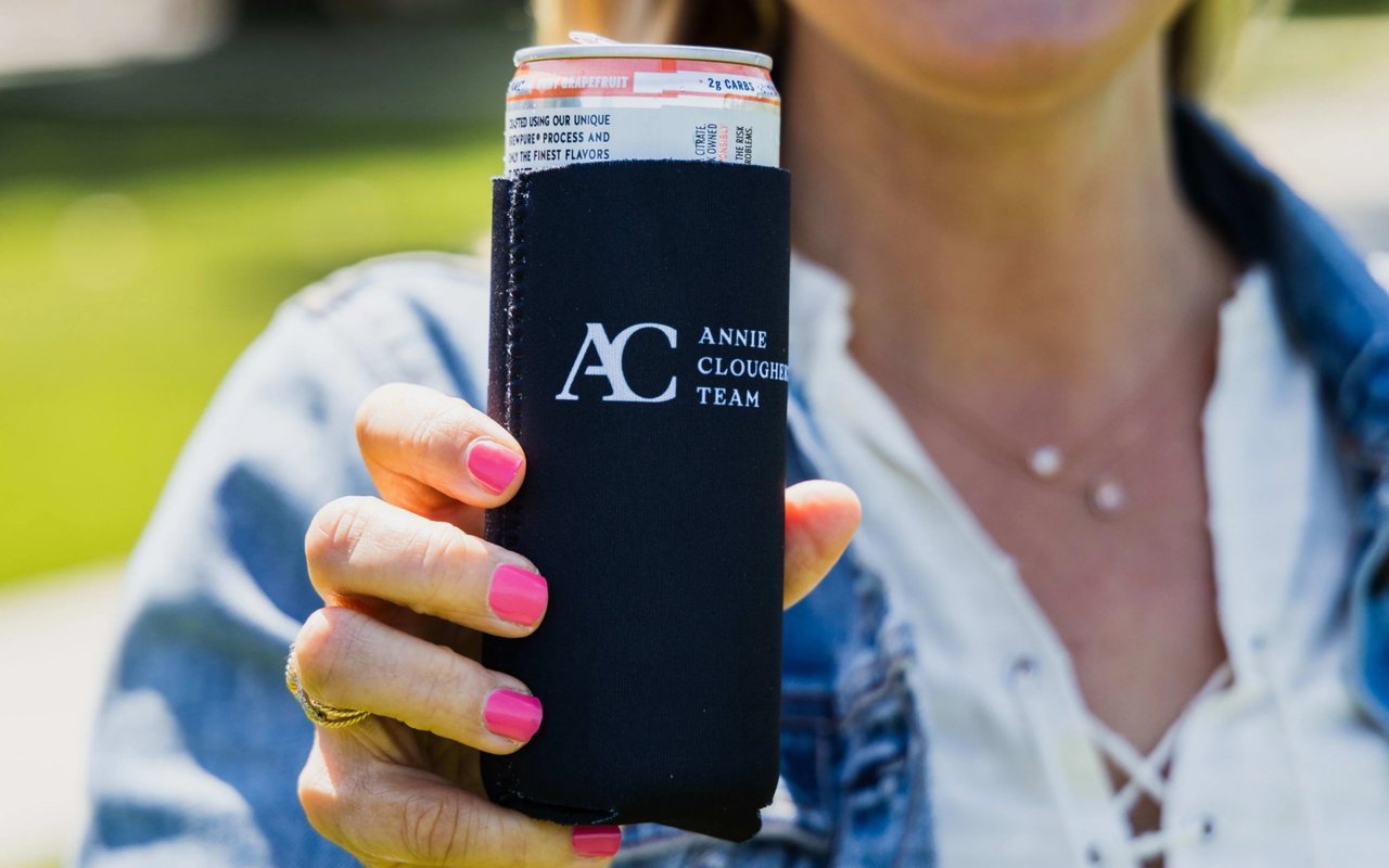 A person with bright pink fingernails holds a black beverage koozie with the logo on it.