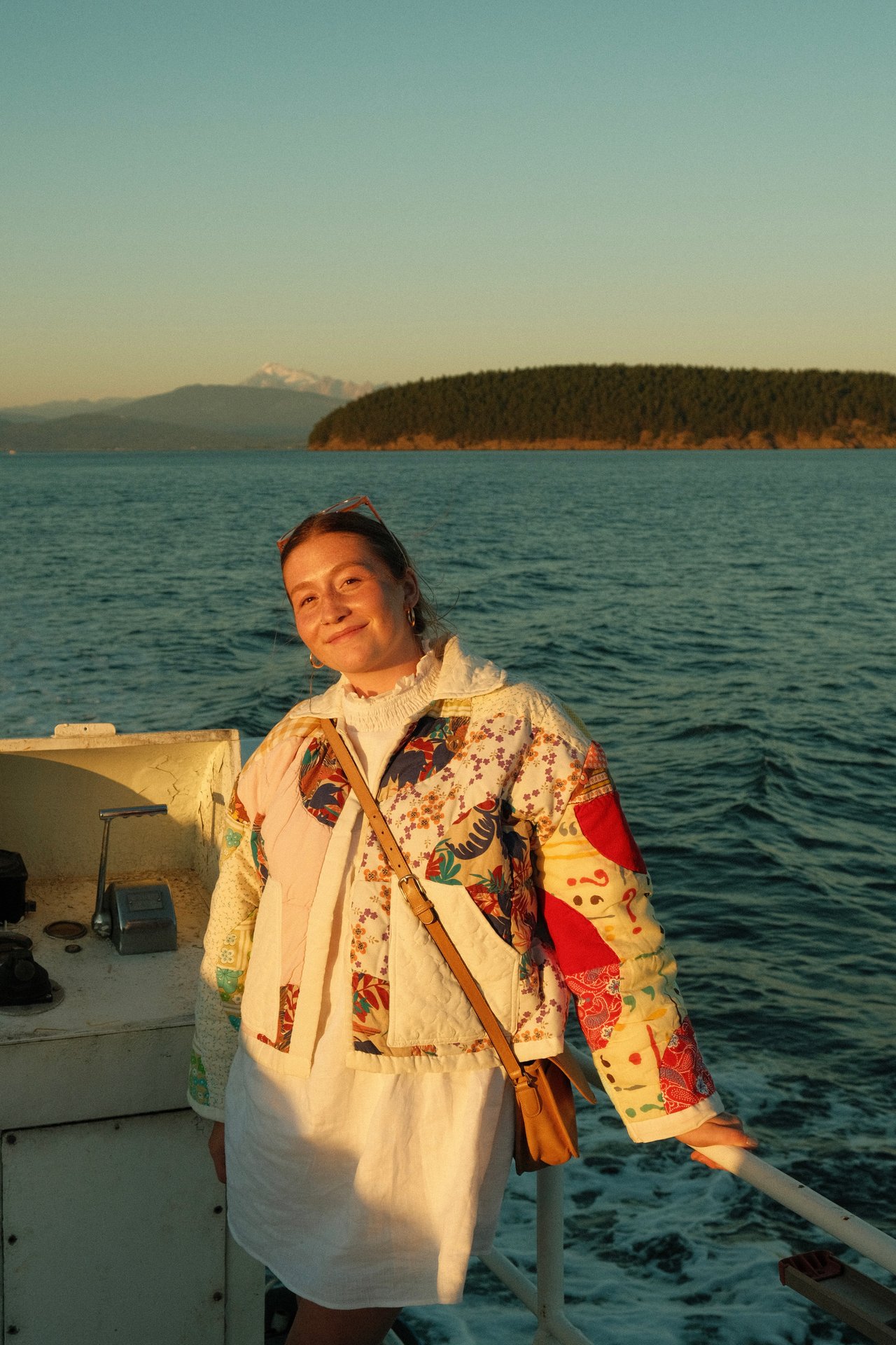 Woman in a colorful patchwork jacket aboard a boat, with water and a forested island behind her.