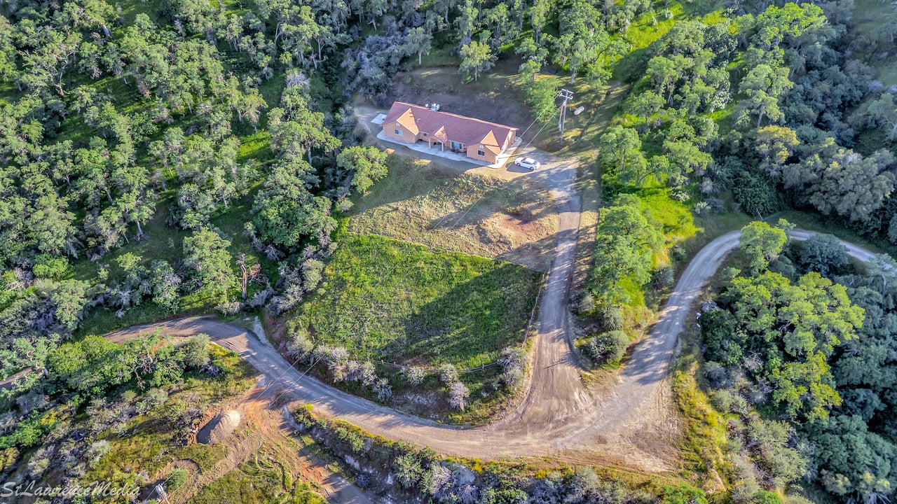 Aerial view of a single-story home amid dense trees, open land, and a winding dirt driveway.