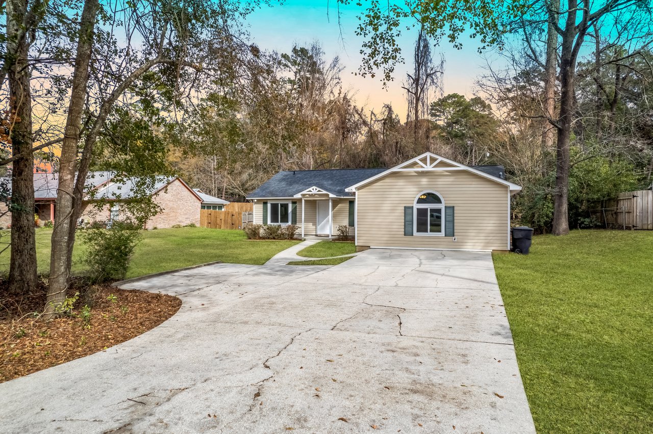 Small beige house with a dark roof, set against a backdrop of trees. A wide driveway leads to the front porch. Tranquil, suburban setting.