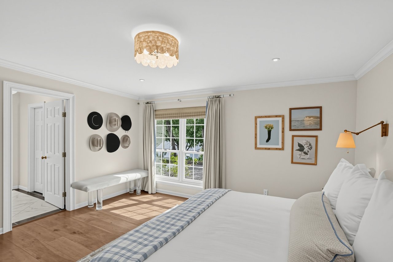 Furnished bedroom with a large white bed, wood floors, and a sunlit window with neutral curtains.