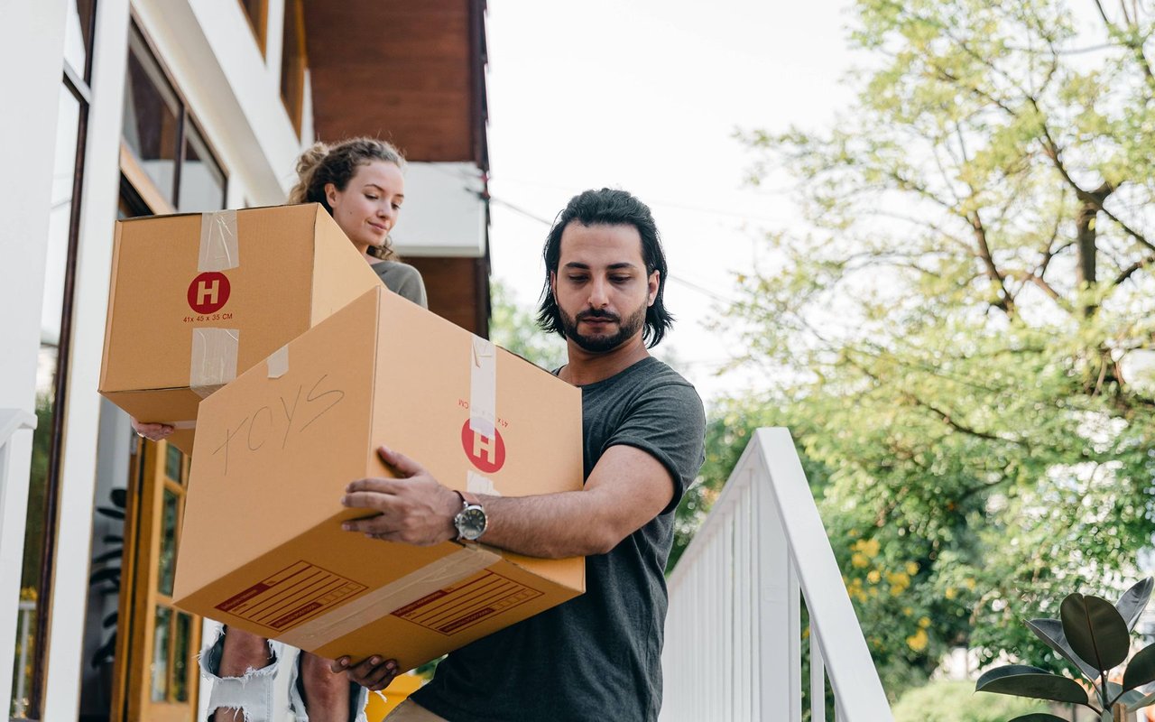A couple carries moving boxes outside a home; the large box in front is labeled “TOYS.”