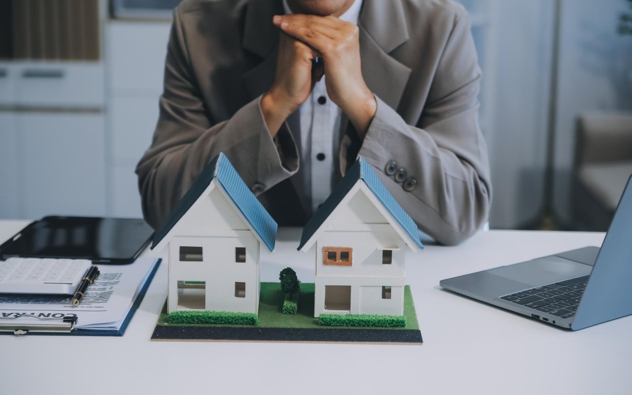 A concerned person considers two model houses at a desk with paperwork and a laptop.