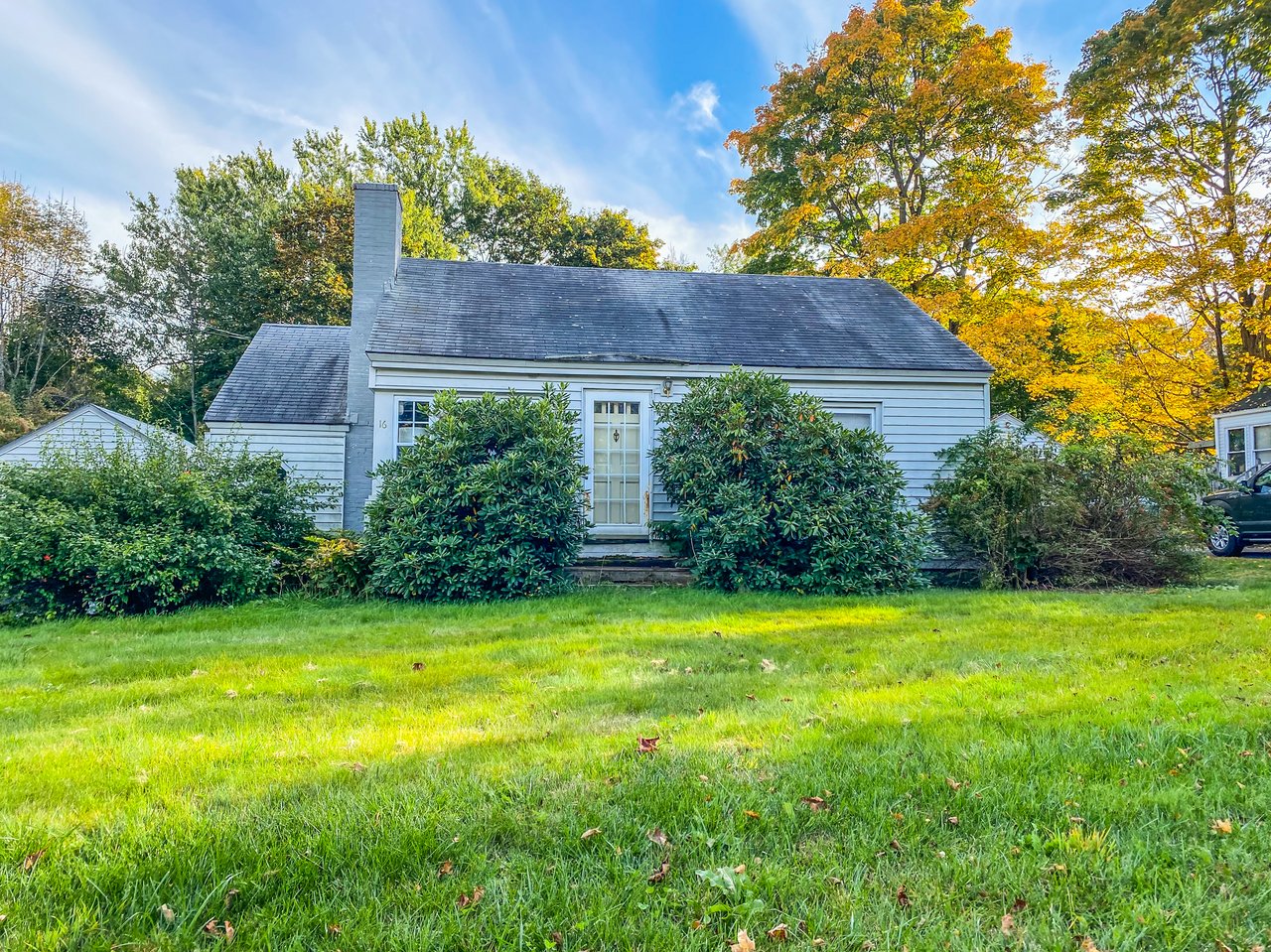 White single-story house with a gray roof, chimney, front shrubs, and a broad lawn.