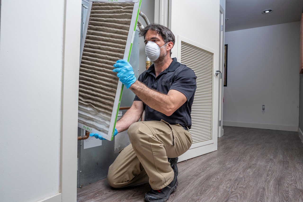 A person wearing a mask and gloves changes a dirty air filter in a residential HVAC system, demonstrating the attention to detail real estate agents value when prepping homes for success.