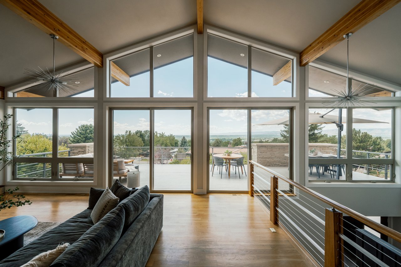 Living room with vaulted wood-beam ceiling and expansive windows overlooking a furnished deck and distant hills.