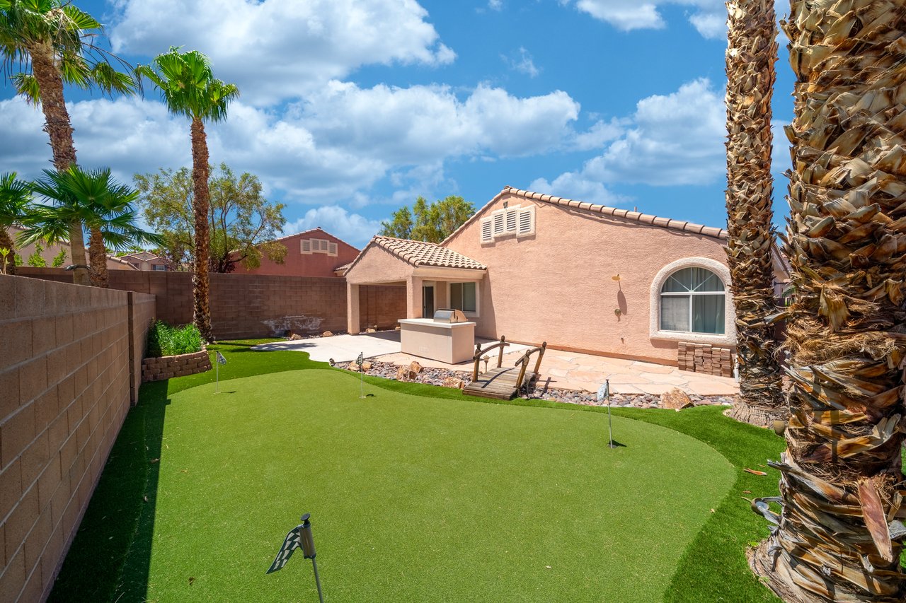 Backyard putting green beside a stucco home, with palm trees and a paved patio.