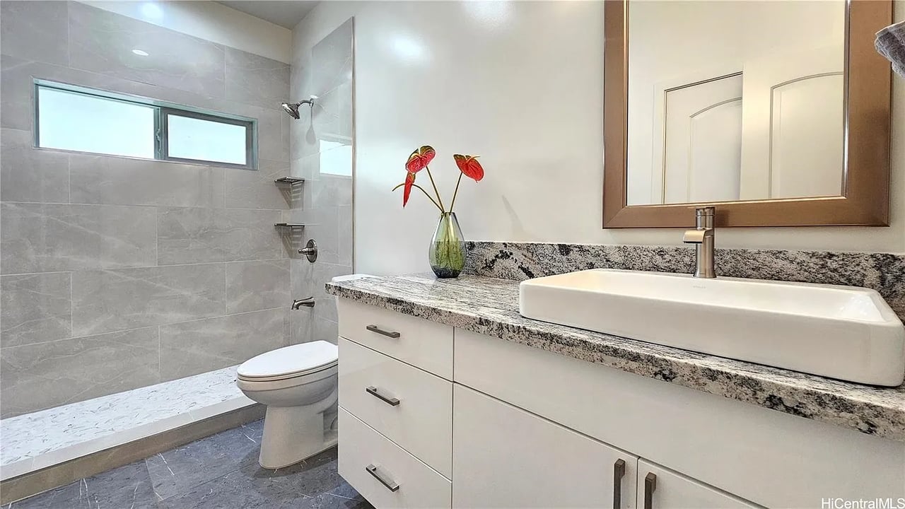 Bathroom with a gray-tiled walk-in shower, high window, and white vanity with granite countertop.