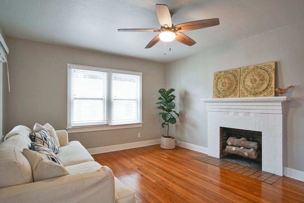 A cozy living room with a white sofa and geometric cushions on a wooden floor. A white brick fireplace with ornate decor and a potted plant add elegance. A ceiling fan brightens the room.