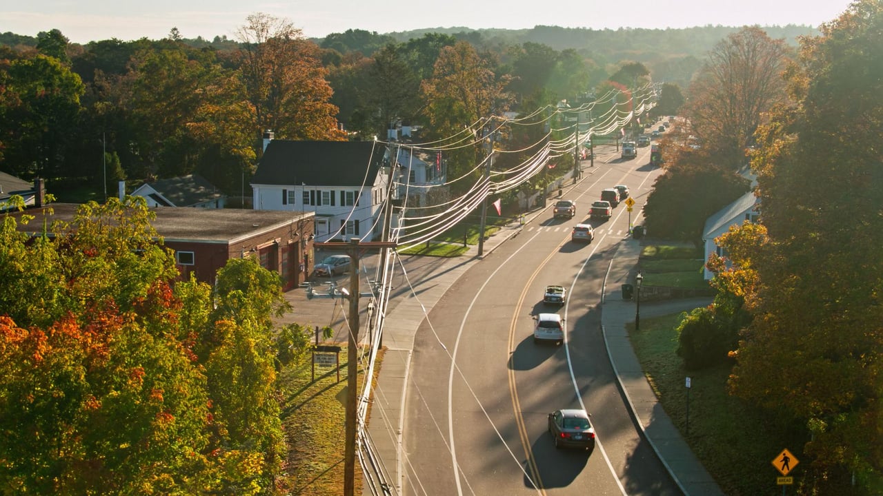 Town road curves past autumn-colored trees and buildings, with wooded hills in the distance.