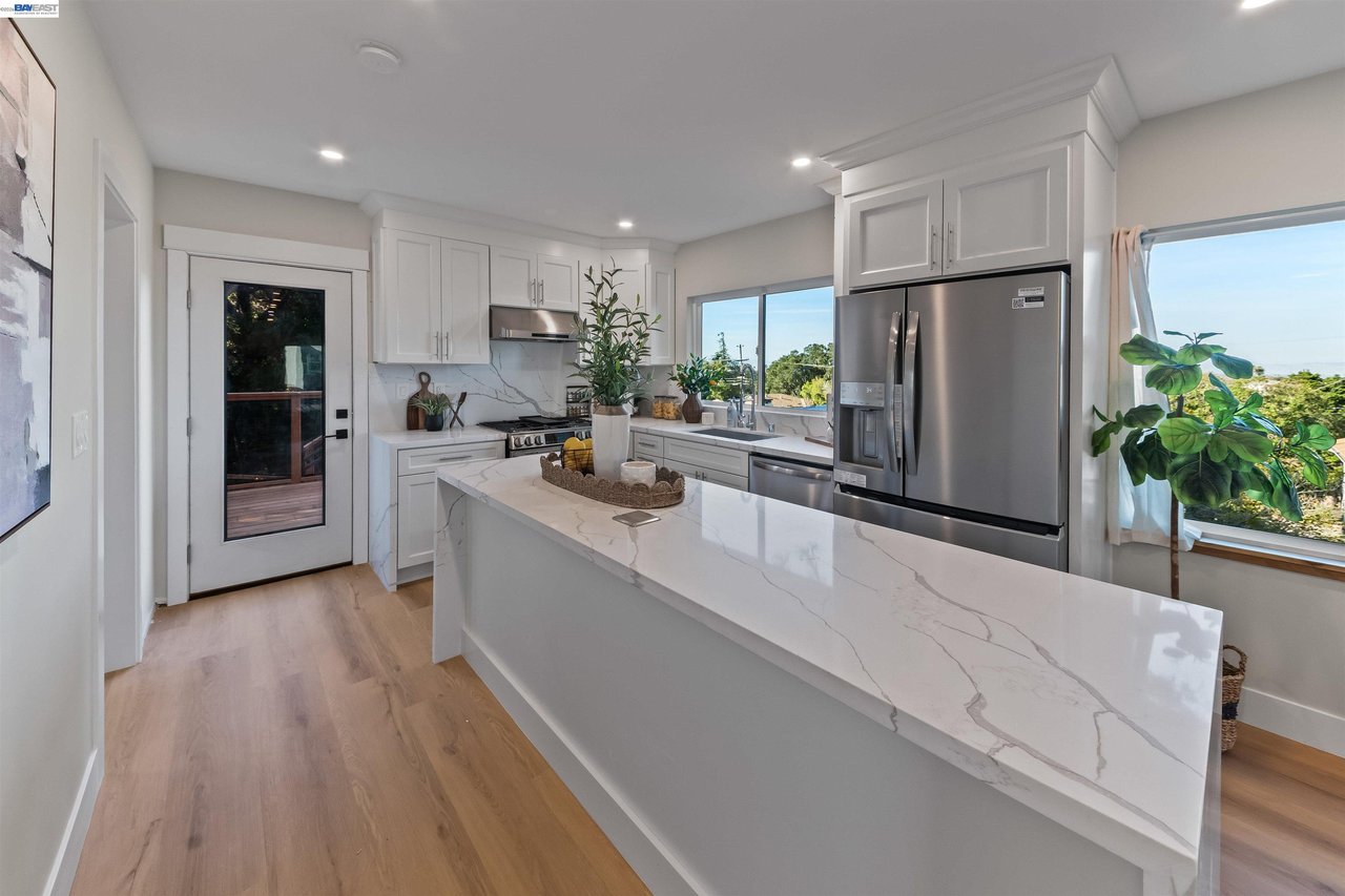 Kitchen with a large veined island, white cabinets, stainless steel appliances, and leafy views through the windows.