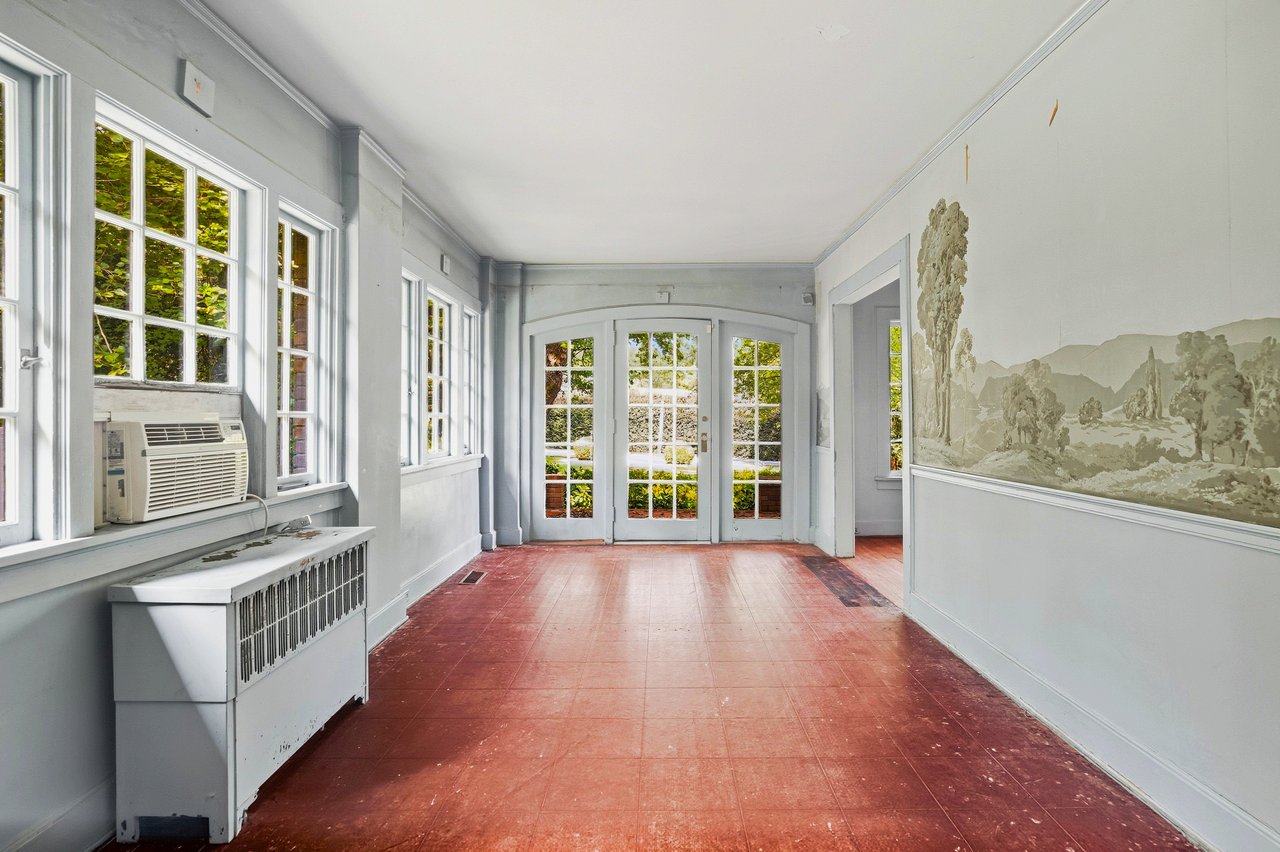 Empty sunroom with red tile floor, tall white-framed windows, and glass doors opening toward greenery.