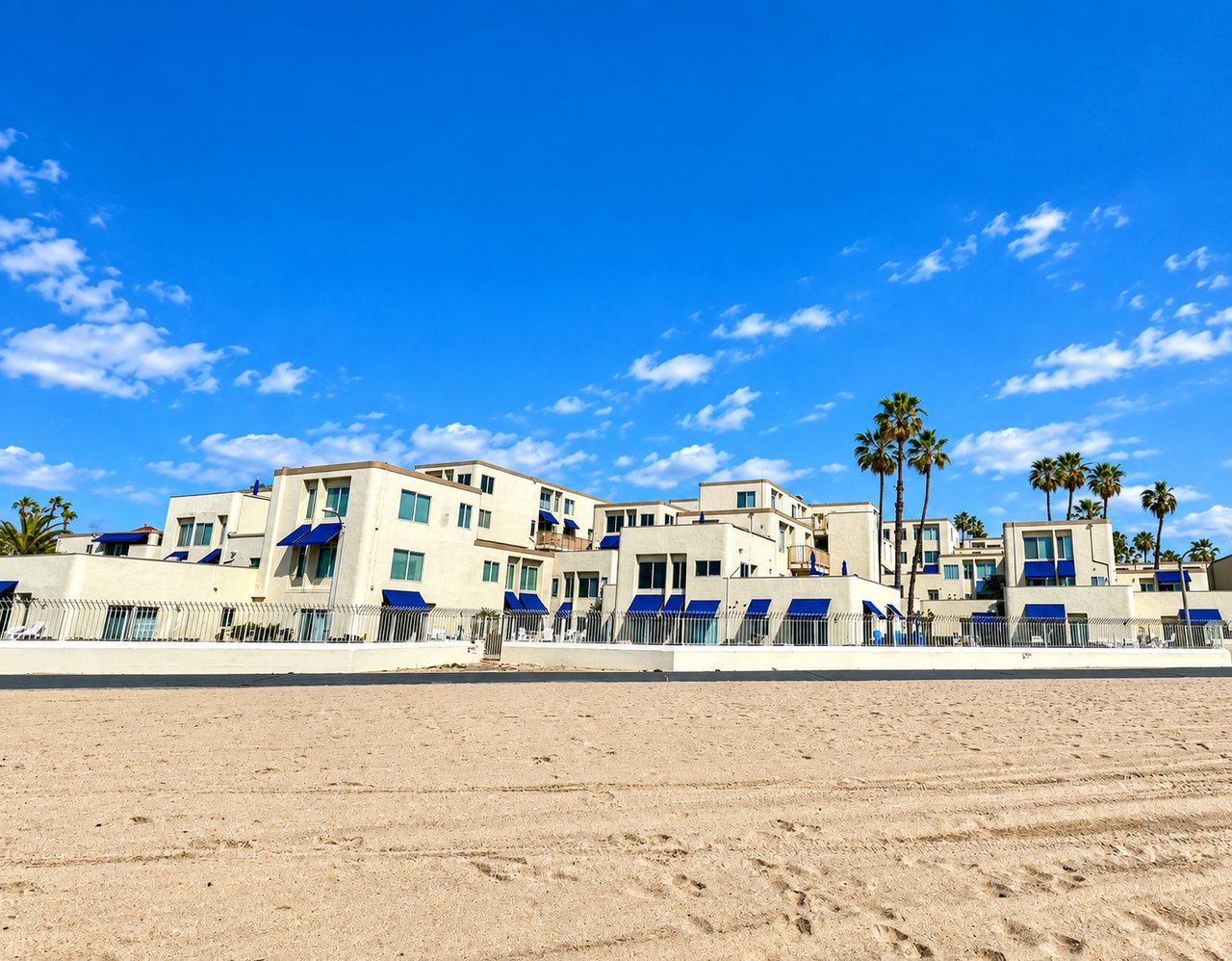 White beachfront condominiums with blue awnings and palm trees behind a sandy beach.