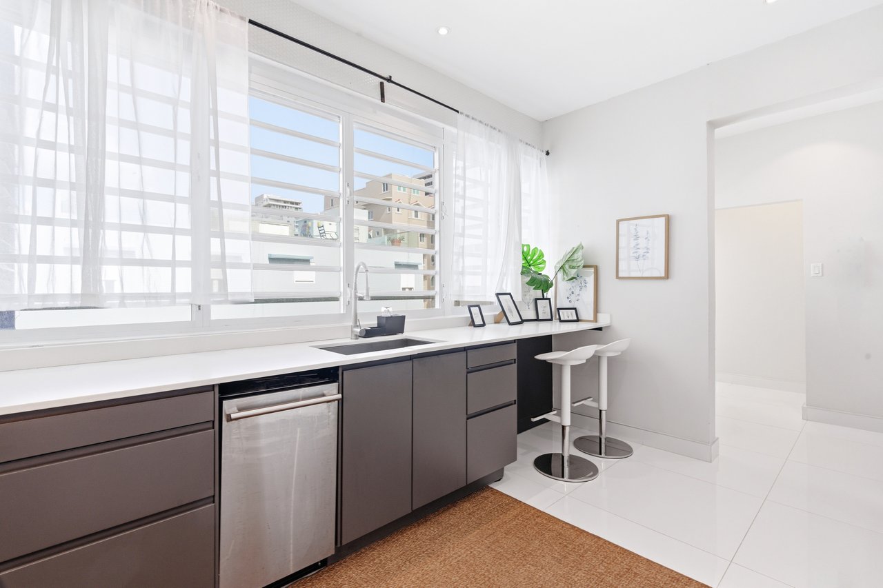 Bright modern kitchen with dark cabinets, a long white counter, large windows, and two bar stools.
