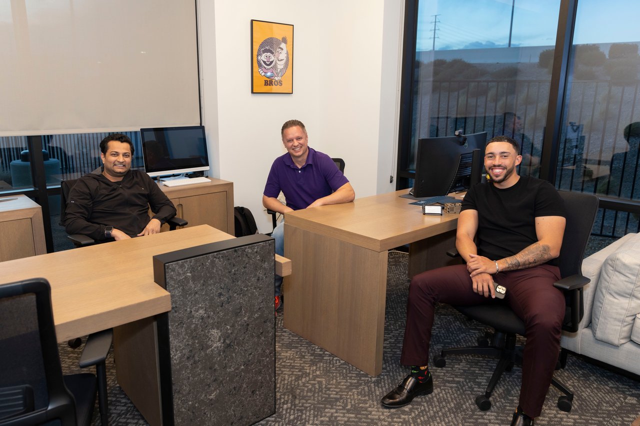 Three men seated behind desks in a modern office with large windows.
