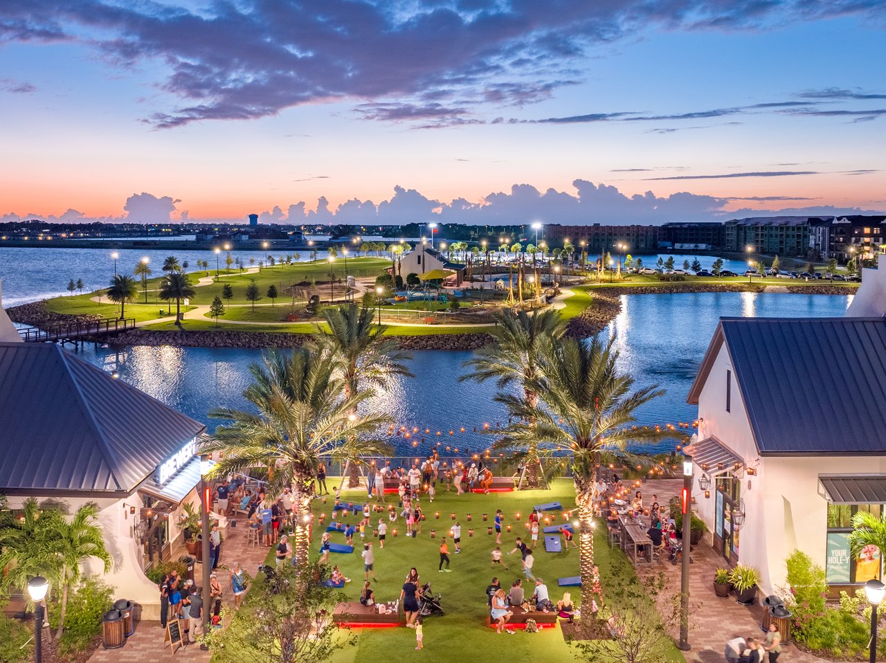 Waterside Place at dusk, with waterfront shops, palm trees, and people gathered on the lawn.