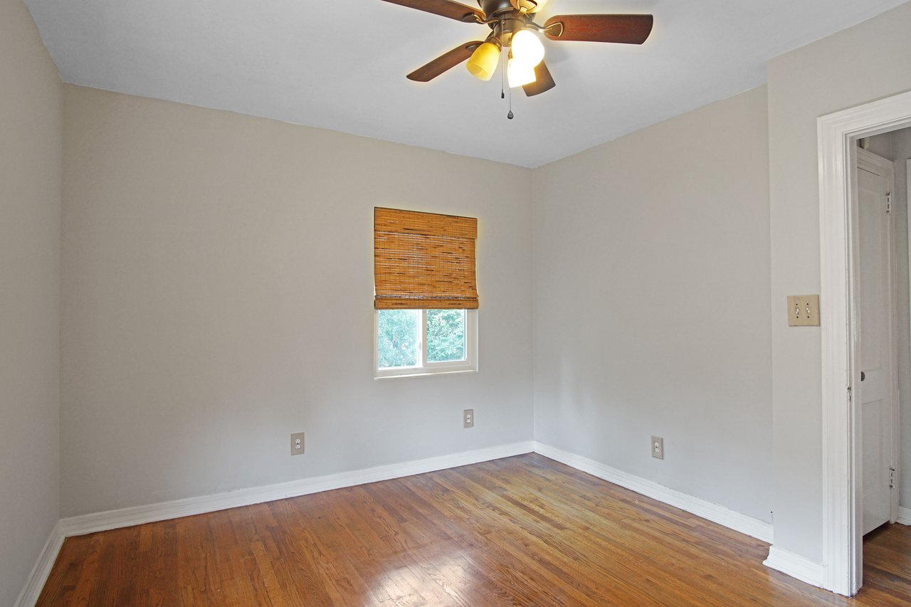 Empty room with light gray walls, wooden floor, ceiling fan with lights, and a small window with bamboo blinds. Simple and serene ambiance.