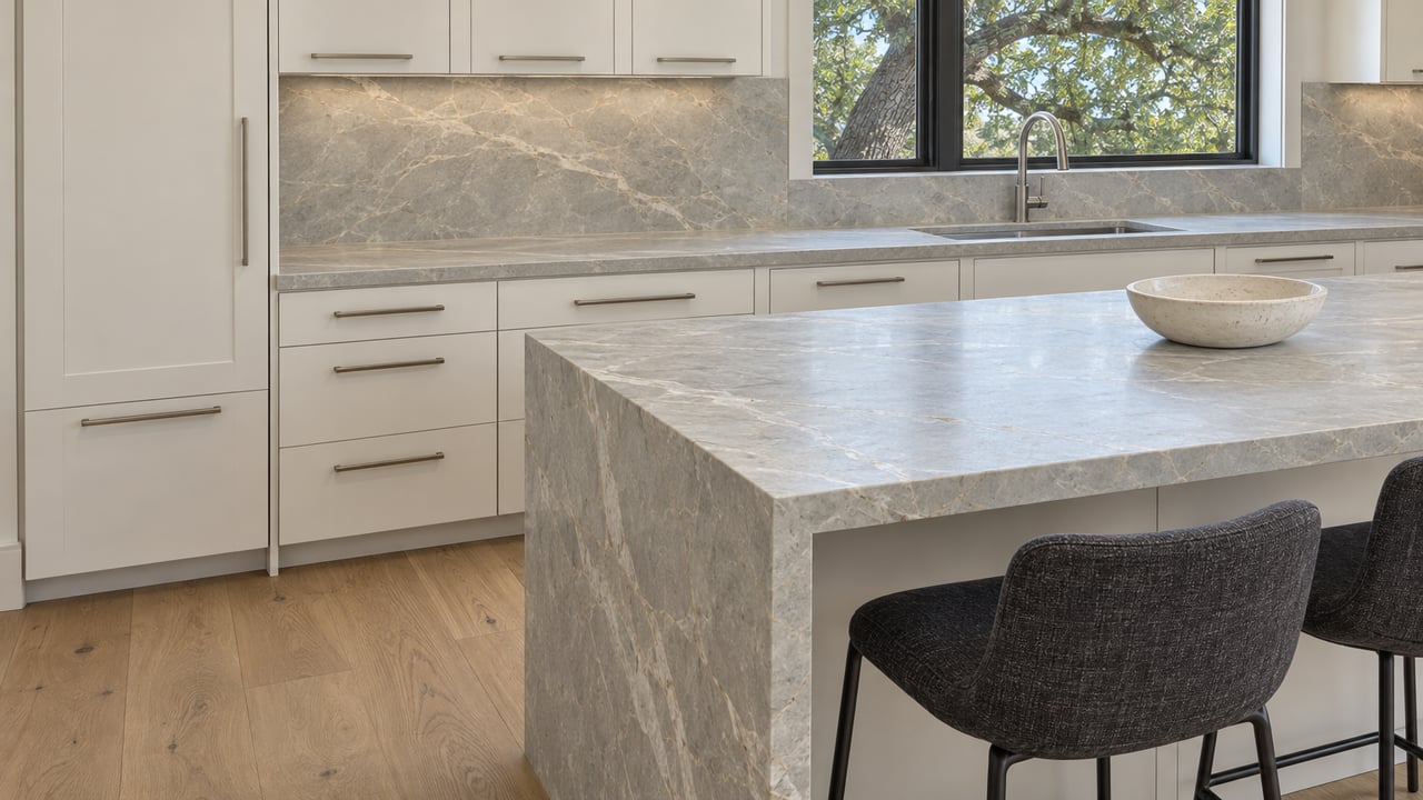 Honed stone waterfall island and two charcoal stools in a bright kitchen with warm-white cabinetry, oak floors and a closed black-framed window.