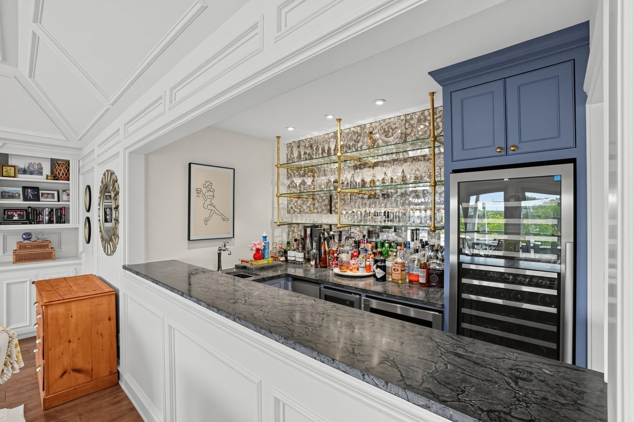 Home bar with a long marble counter, brass glass shelves, and blue cabinetry with a wine refrigerator.