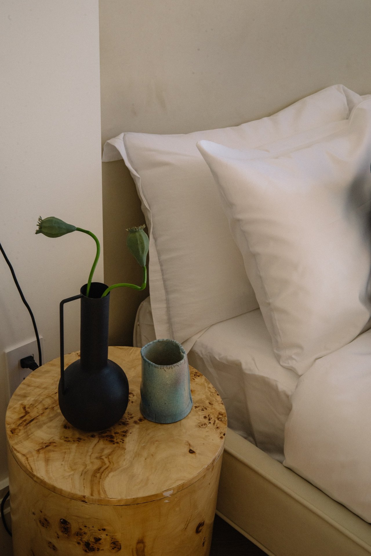Bedroom pillows beside a round burl wood bedside table with a vase and ceramic cup.