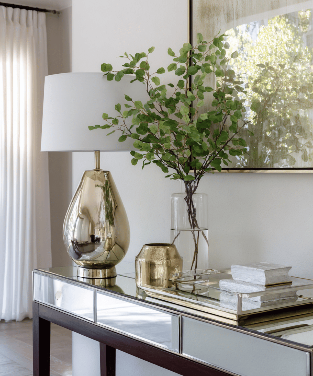 Mirrored console table styled with a gold lamp, leafy branches, and decorative objects beside a bright window.