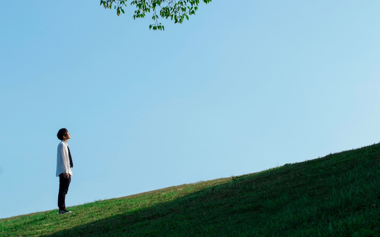A person in business attire looks upward from a grassy hillside beneath a wide blue sky.