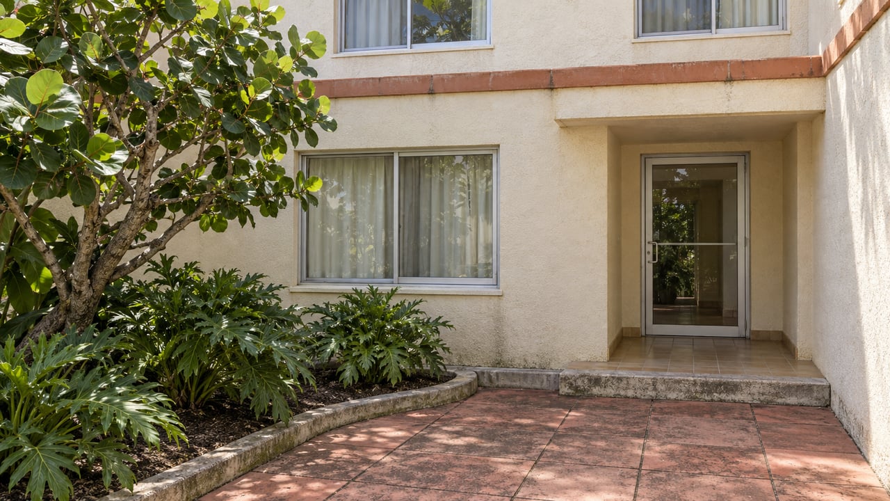 Cream stucco condominium entrance with aluminum-framed windows, terracotta coping and a planted coral-colored courtyard.