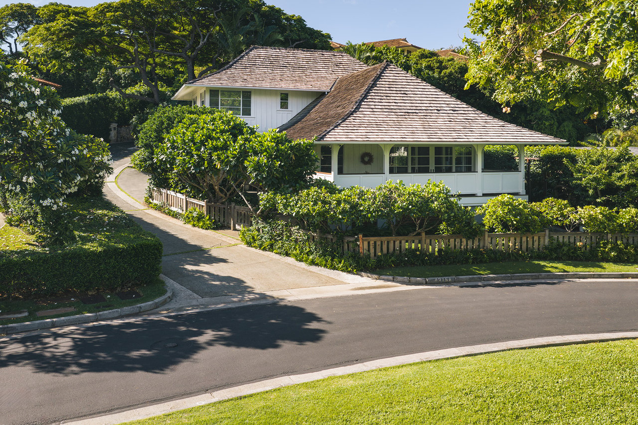 White two-story home with broad shingled roofs, a covered veranda, and lush landscaping beside a curved driveway.