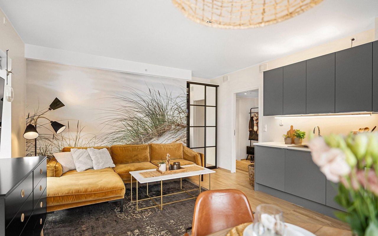 Open-plan living room with a mustard sectional, botanical mural, and dark kitchen cabinetry.
