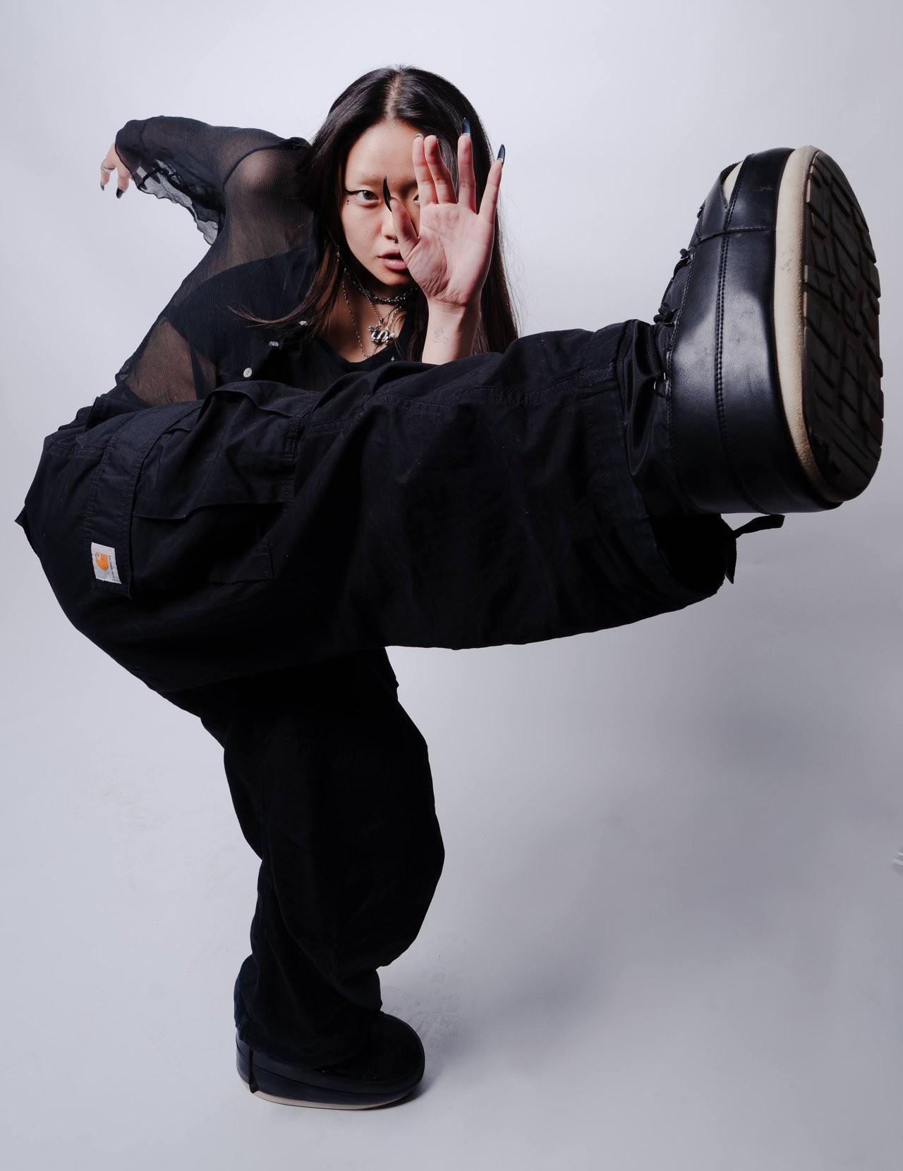 Dancer Jin poses in black streetwear, balancing with one boot sole facing the camera.
