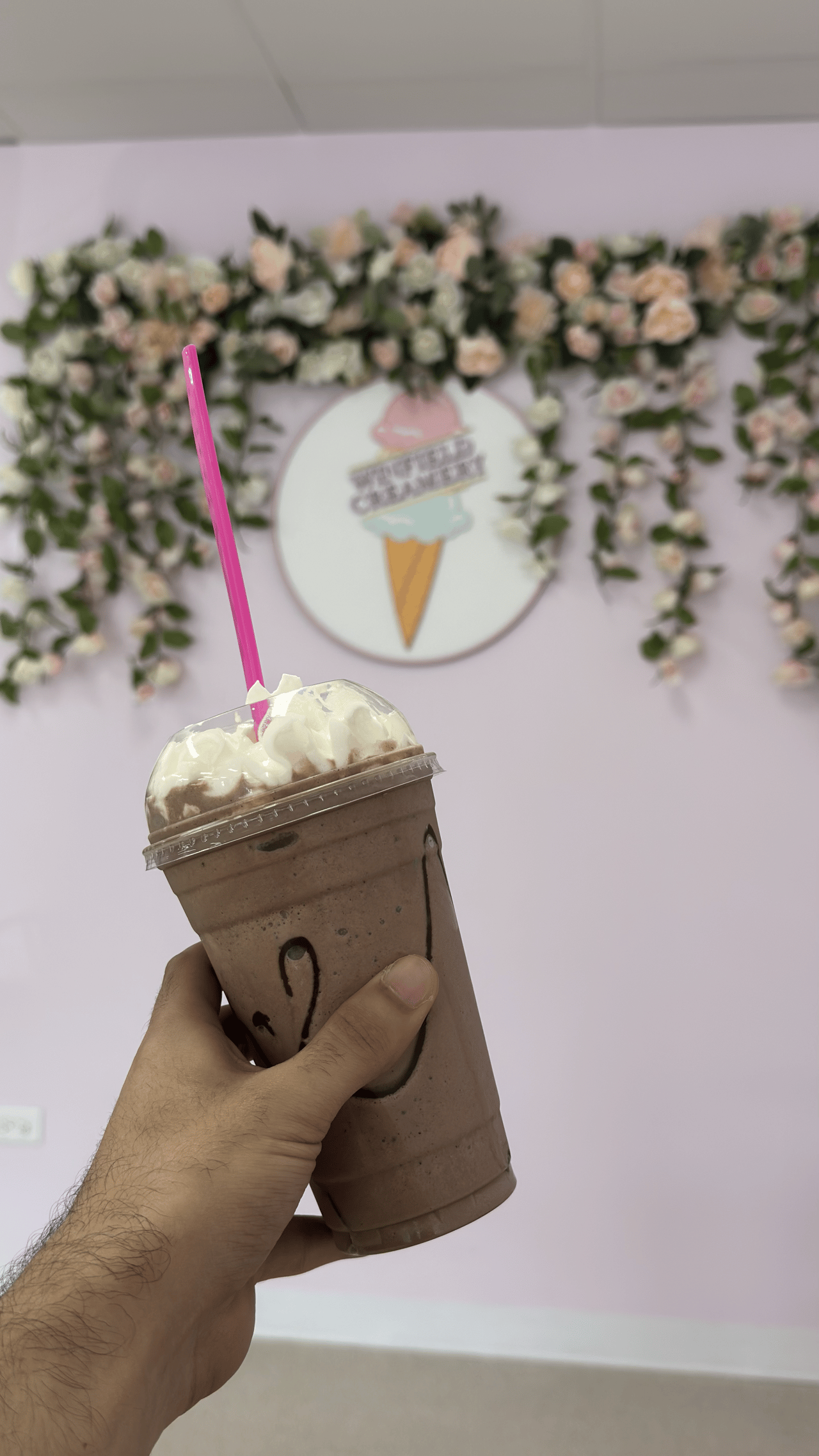 Hand holding a clear plastic cup filled with a chocolate milkshake topped with whipped cream and a bright pink straw, set against a floral wall with the Winfield Creamery logo.