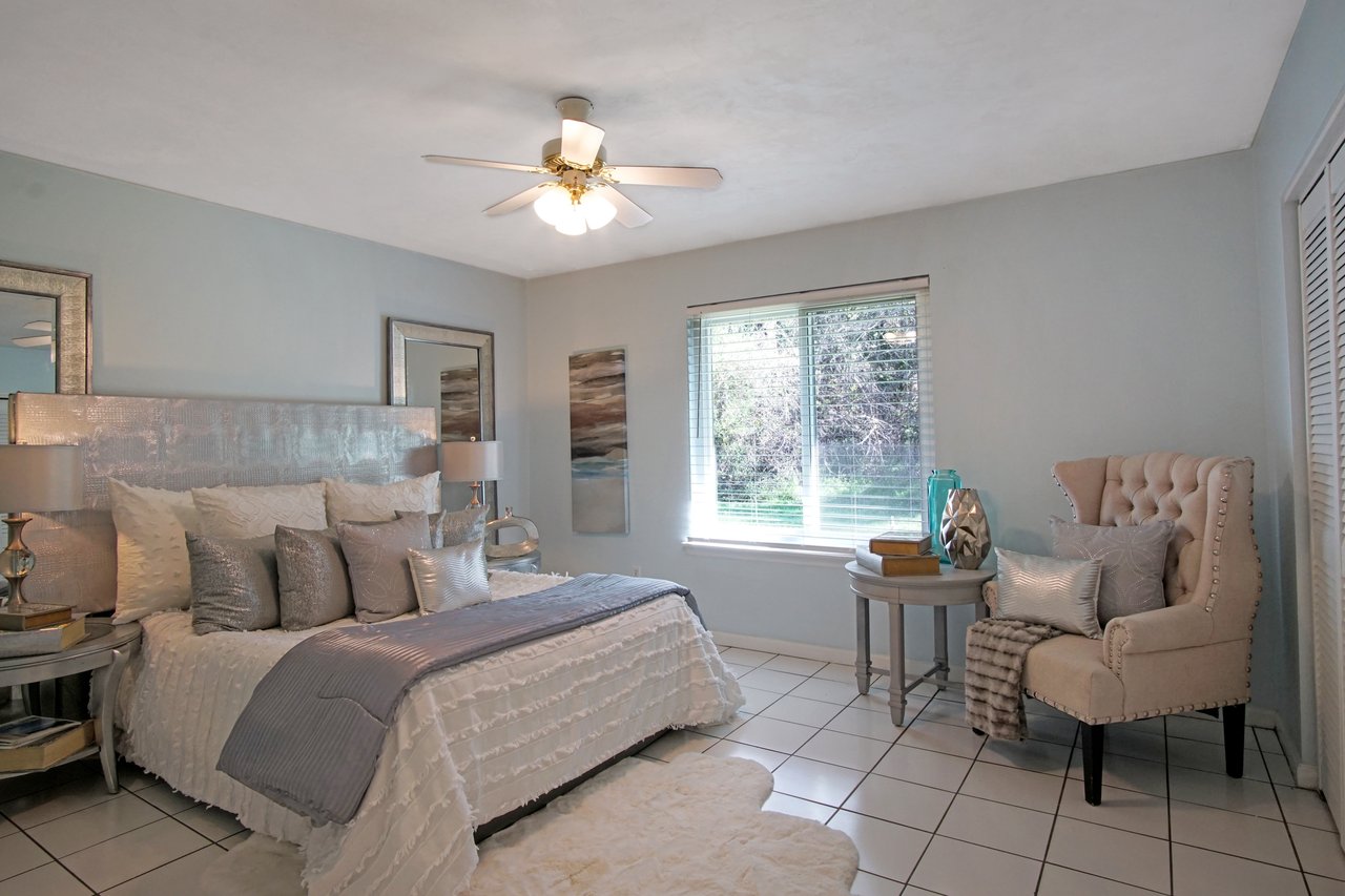 Elegant bedroom with a king-size bed adorned with white and silver pillows and a soft throw. A cozy armchair and side table sit by a window, emitting natural light and a serene ambiance.Elegant bedroom with a king-size bed adorned with white and silver pillows and a soft throw. A cozy armchair and side table sit by a window, emitting natural light and a serene ambiance.