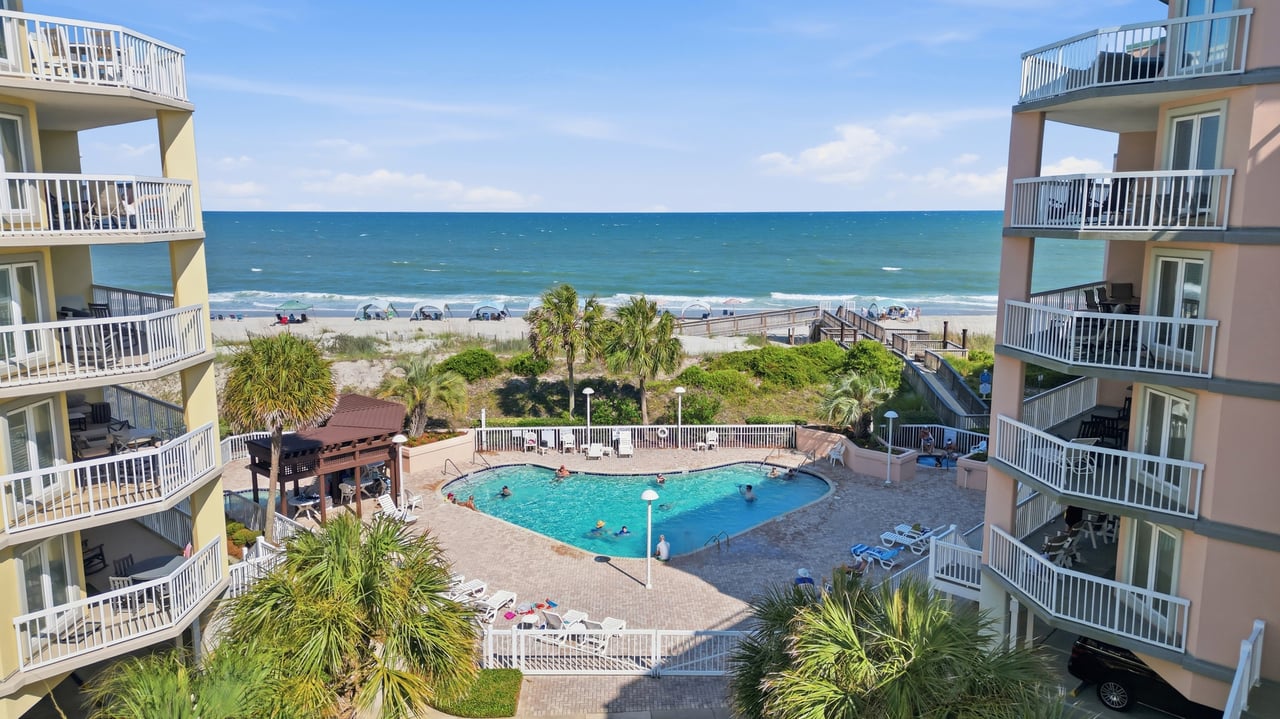 Condominium courtyard pool overlooking a sandy beach and ocean, framed by balcony buildings.