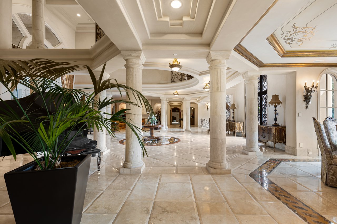 Mansion foyer with marble floors, classical columns, and a large potted palm.